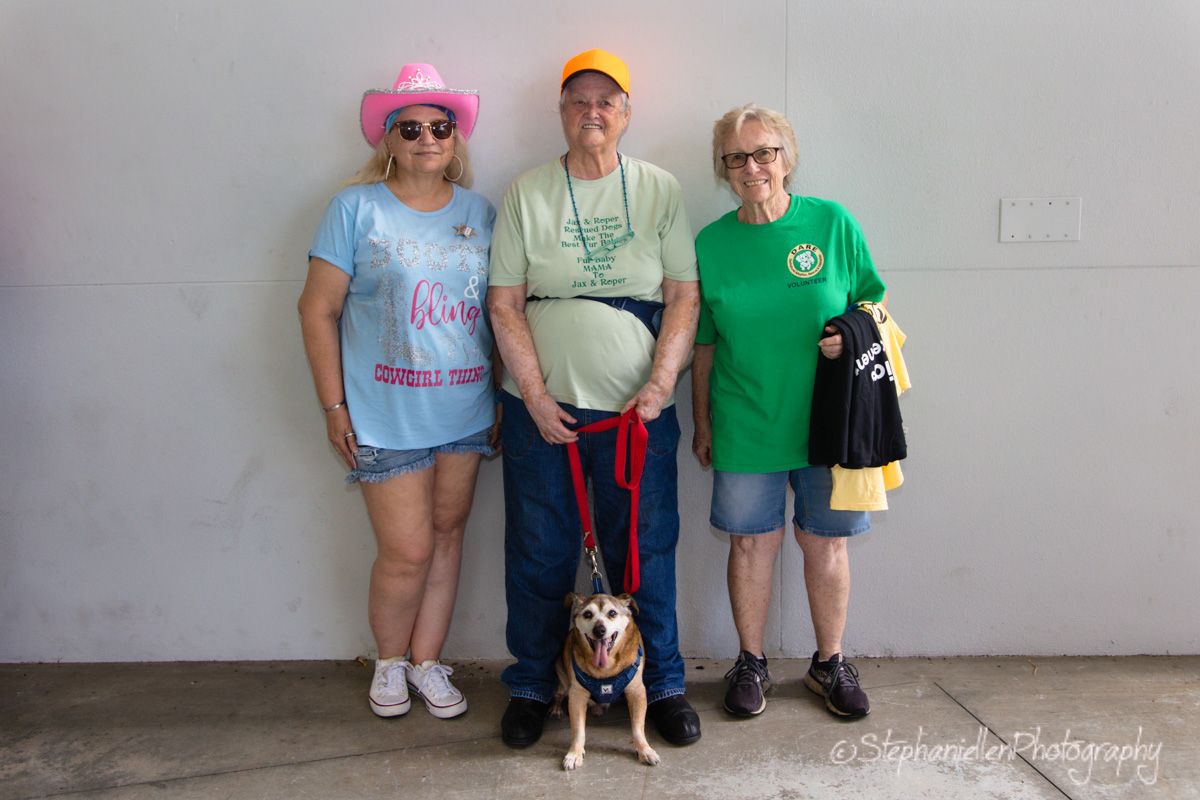 Three people and a dog on a red leash pose together against a gray wall outdoors.