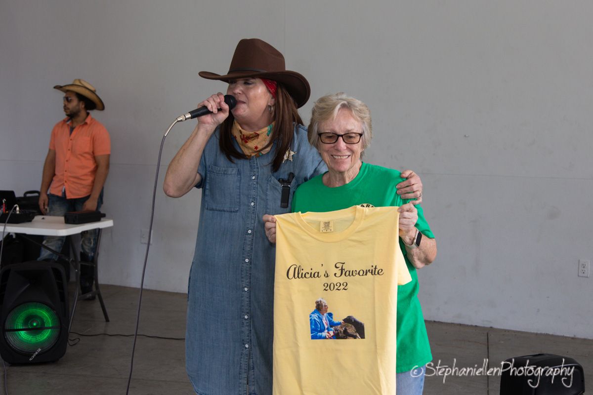 Two women pose together at an outdoor event, one holding a yellow 2022 t-shirt while the other speaks into a microphone.