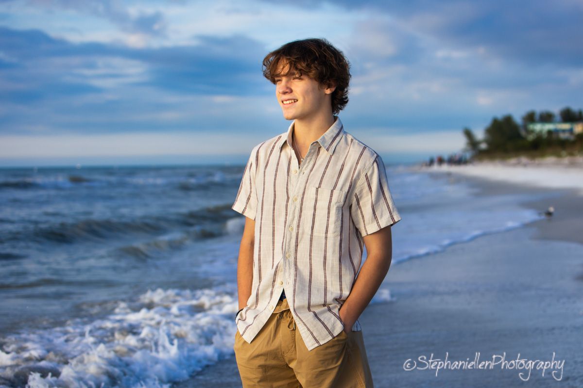 Teen boy in striped shirt standing on beach with ocean waves and dramatic blue sky backdrop.
