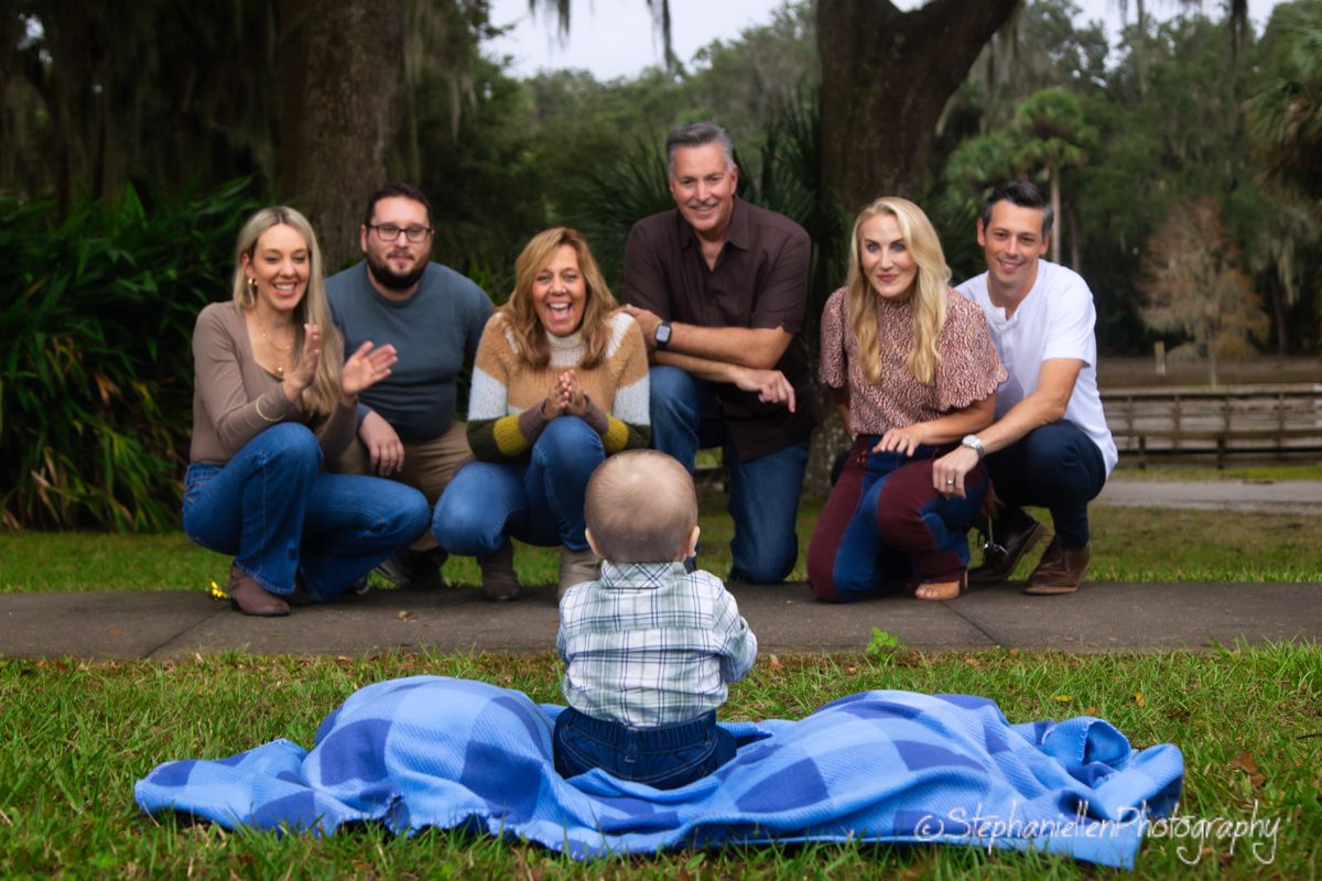 Six adults gathered around a baby sitting on blue blanket in a sunny park.