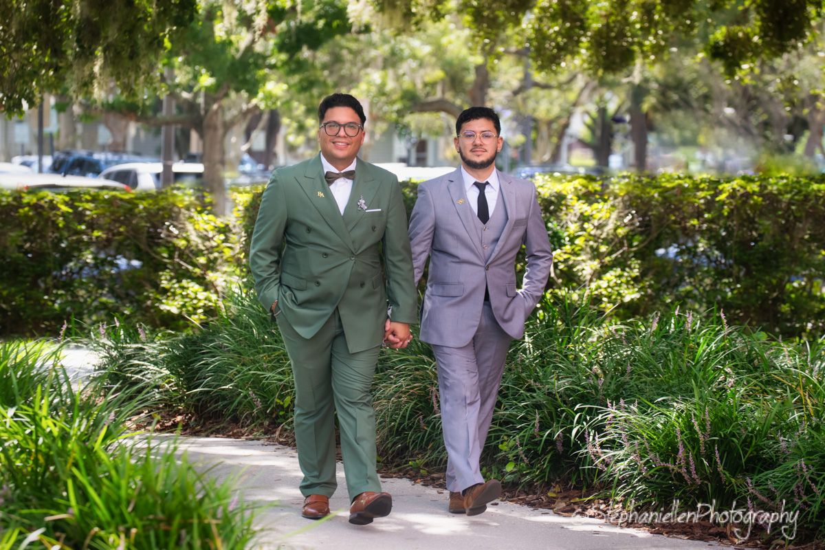 Two grooms in green and gray suits walk hand in hand along a tree-lined path.
