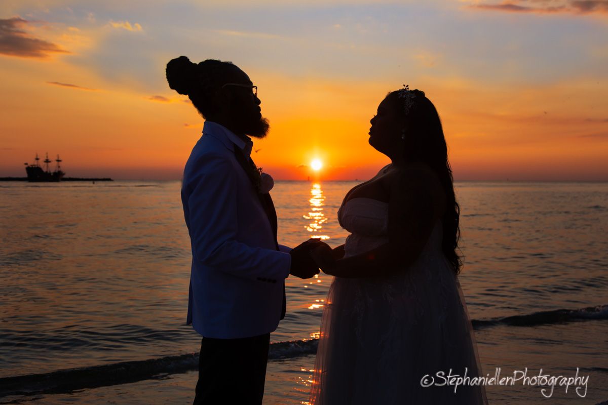Romantic silhouette of bride and groom holding hands on beach at vibrant orange sunset.