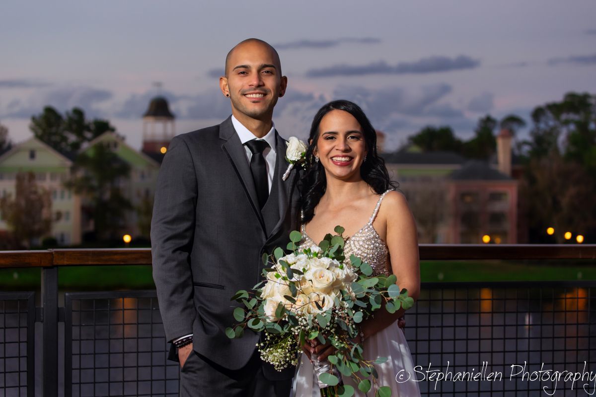 Smiling couple in wedding attire holding floral bouquet on rooftop at dusk.