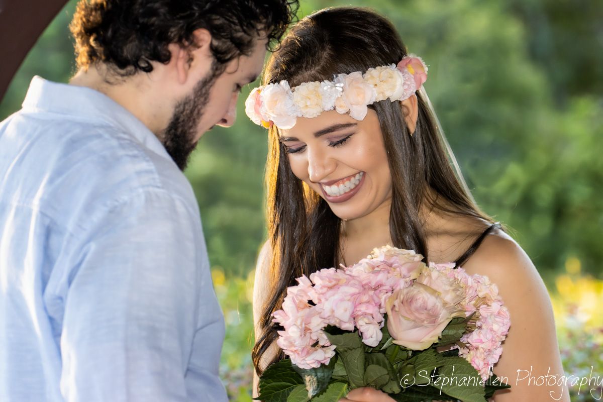 Smiling bride wearing floral crown admires pink bouquet of roses and hydrangeas at outdoor wedding ceremony.