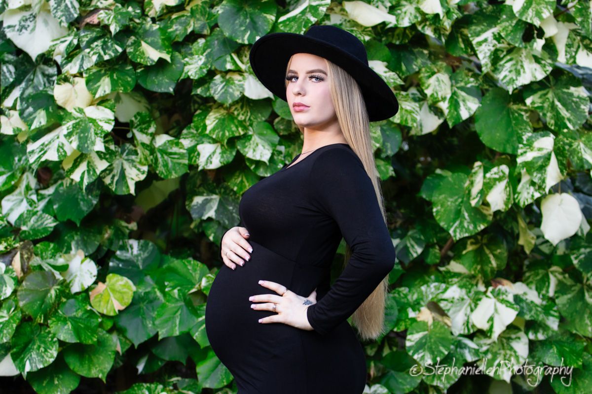 Pregnant woman in black dress and wide-brim hat posing in front of lush green ivy wall.