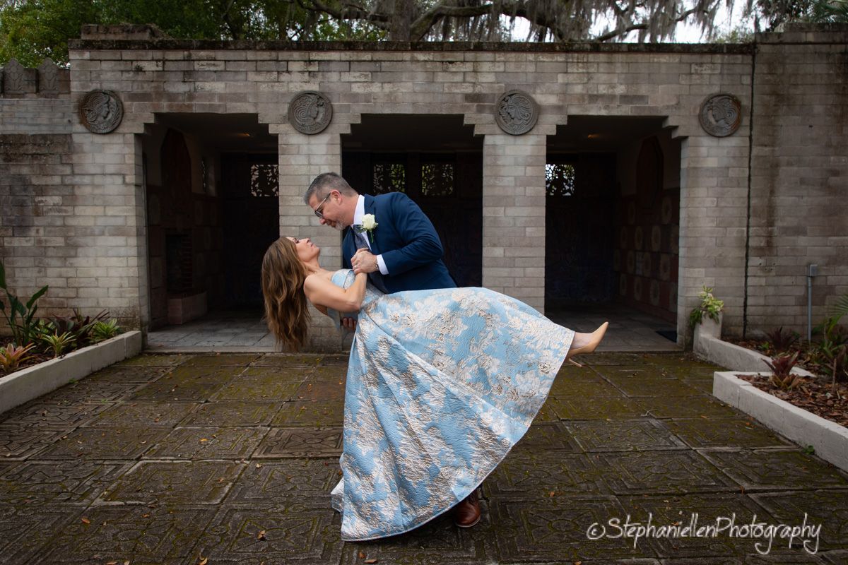 Groom dipping bride in a blue floral dress outside a stone building in a romantic wedding photo.