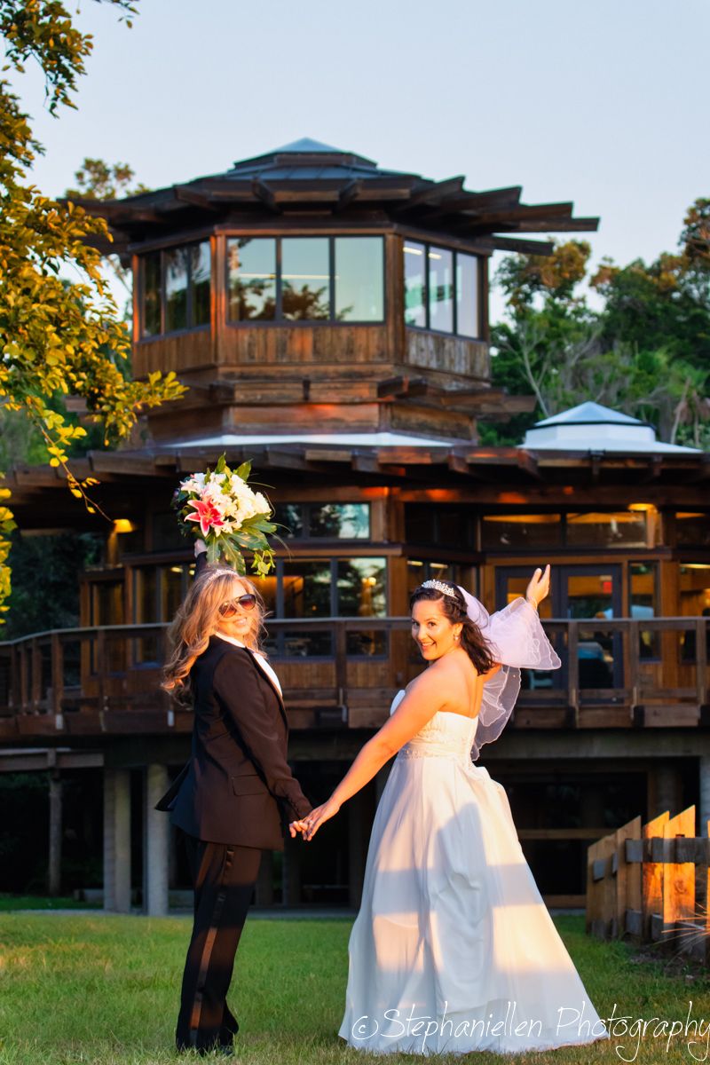 Two brides hold hands on lawn before rustic wooden venue, one in white gown and one in black suit with bouquet.