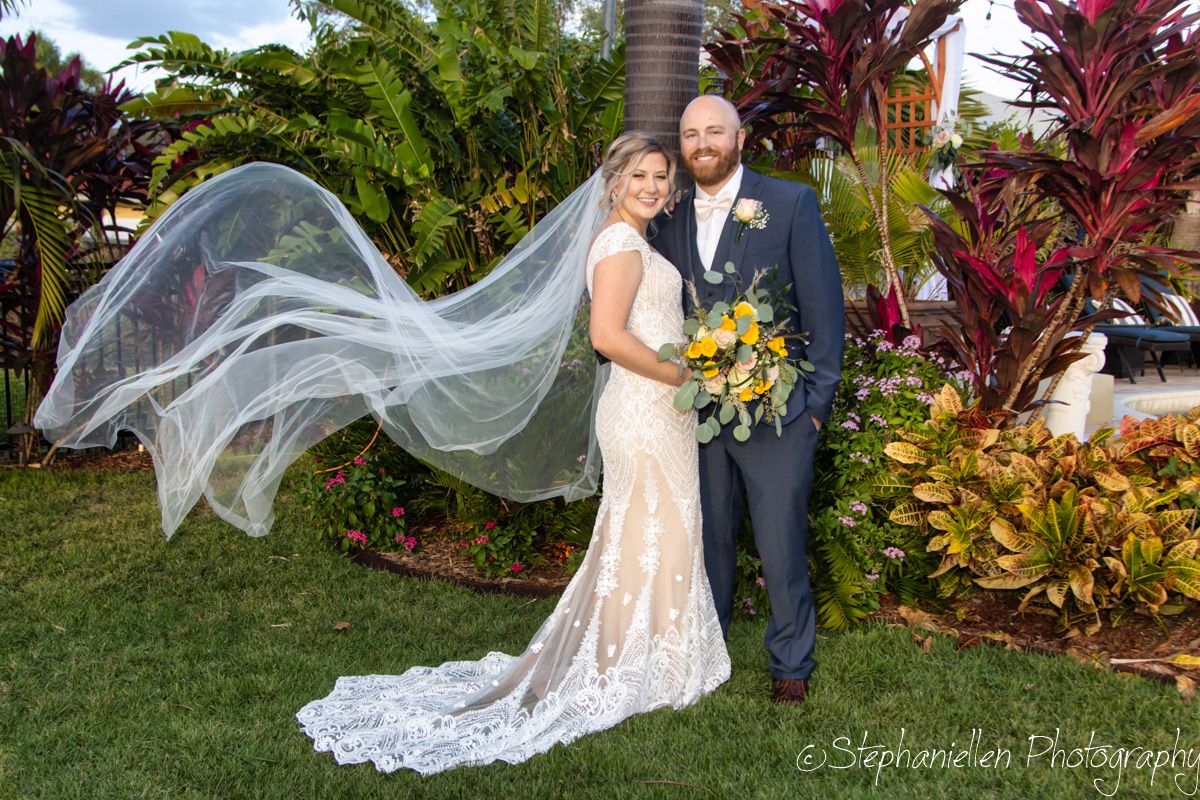 Bride in lace gown with flowing veil billowing in wind stands with groom in navy suit outdoors.