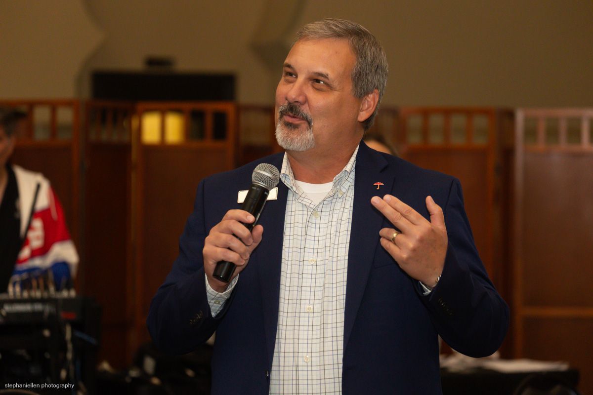 Man in blue blazer speaks into a microphone at an indoor event or conference.