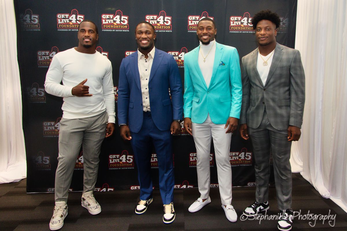 Four well-dressed men pose together on step-and-repeat backdrop at formal charity gala.