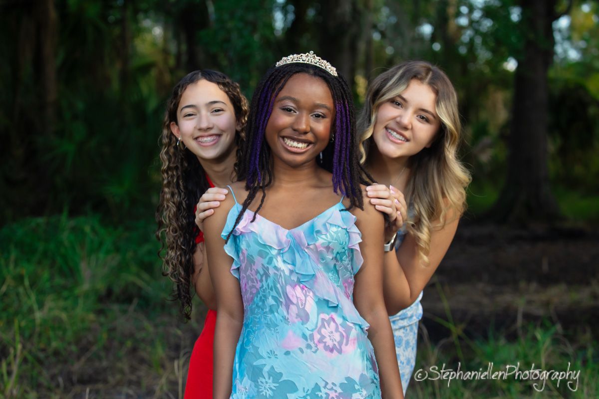 Three teenage girls smiling and posing together outdoors, middle girl wearing a tiara and blue floral dress.
