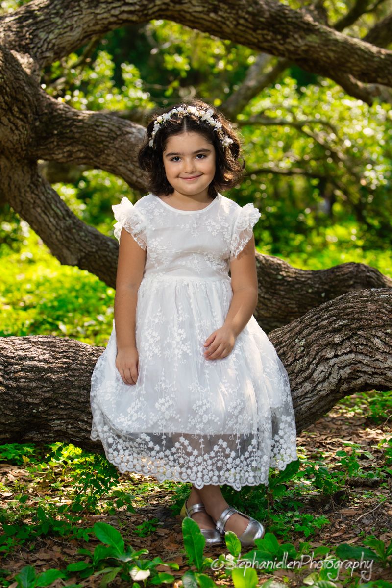 Young girl in white lace dress with floral crown sits on oak tree branch in lush green park setting.