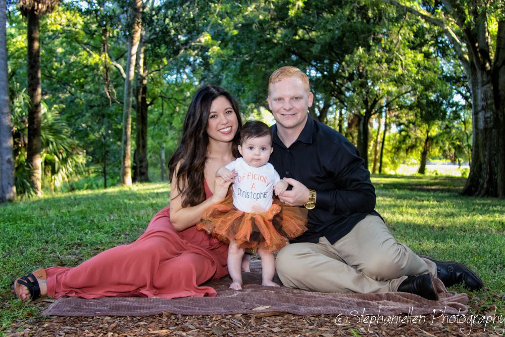 Family portrait of parents with baby girl in orange tutu sitting on blanket in lush green park.
