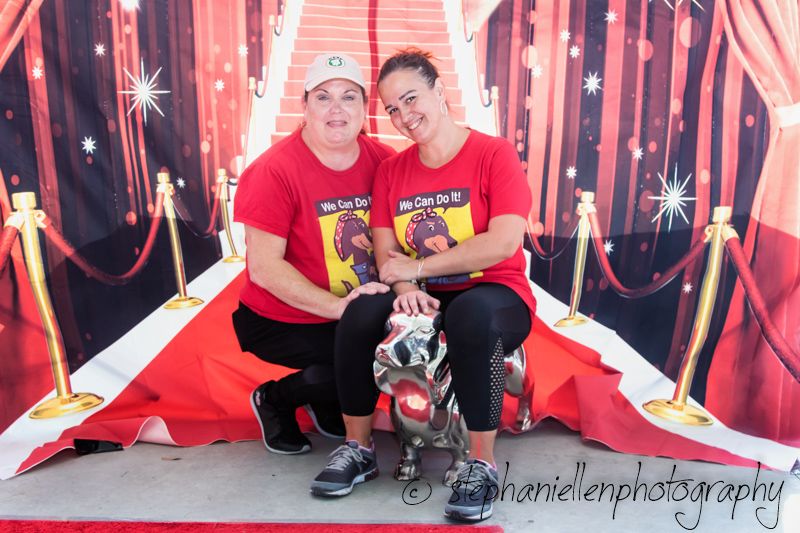 Two women in matching red shirts pose on a red carpet backdrop with a small dog at a themed photography event.
