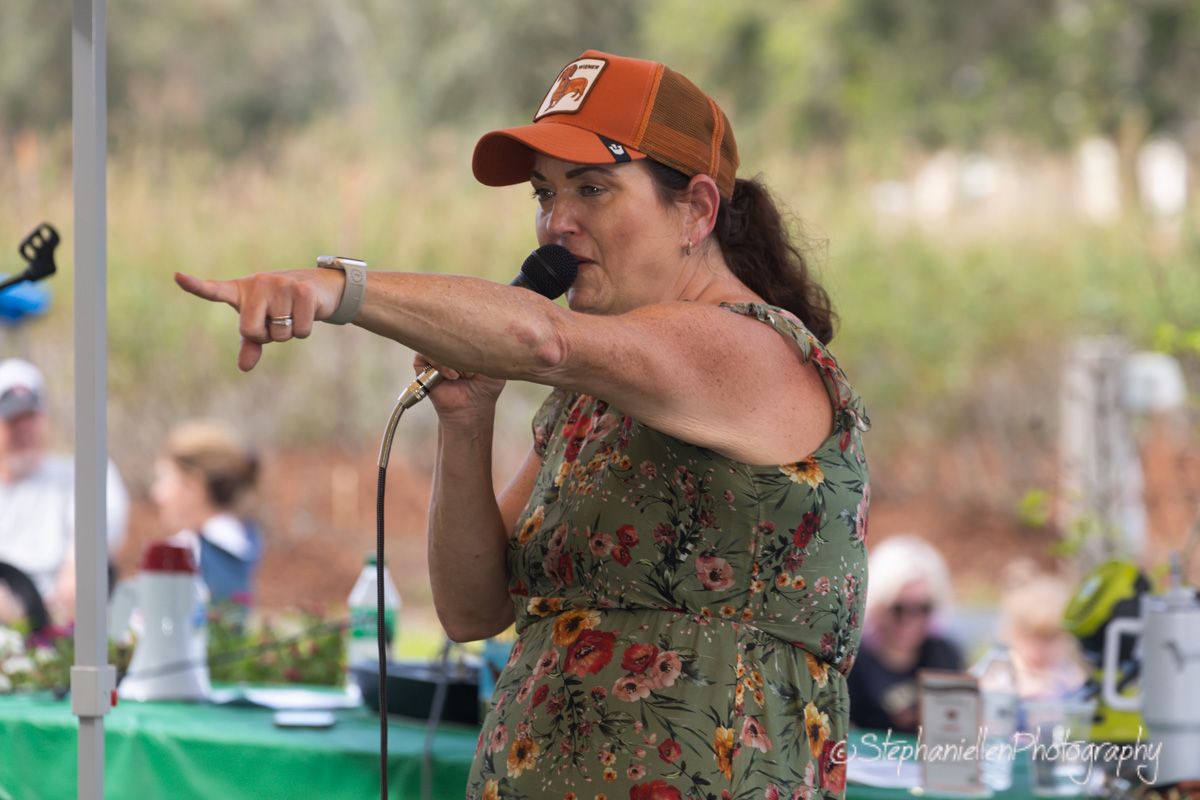 Woman in floral dress and orange cap speaks into microphone at outdoor event, pointing expressively at audience.