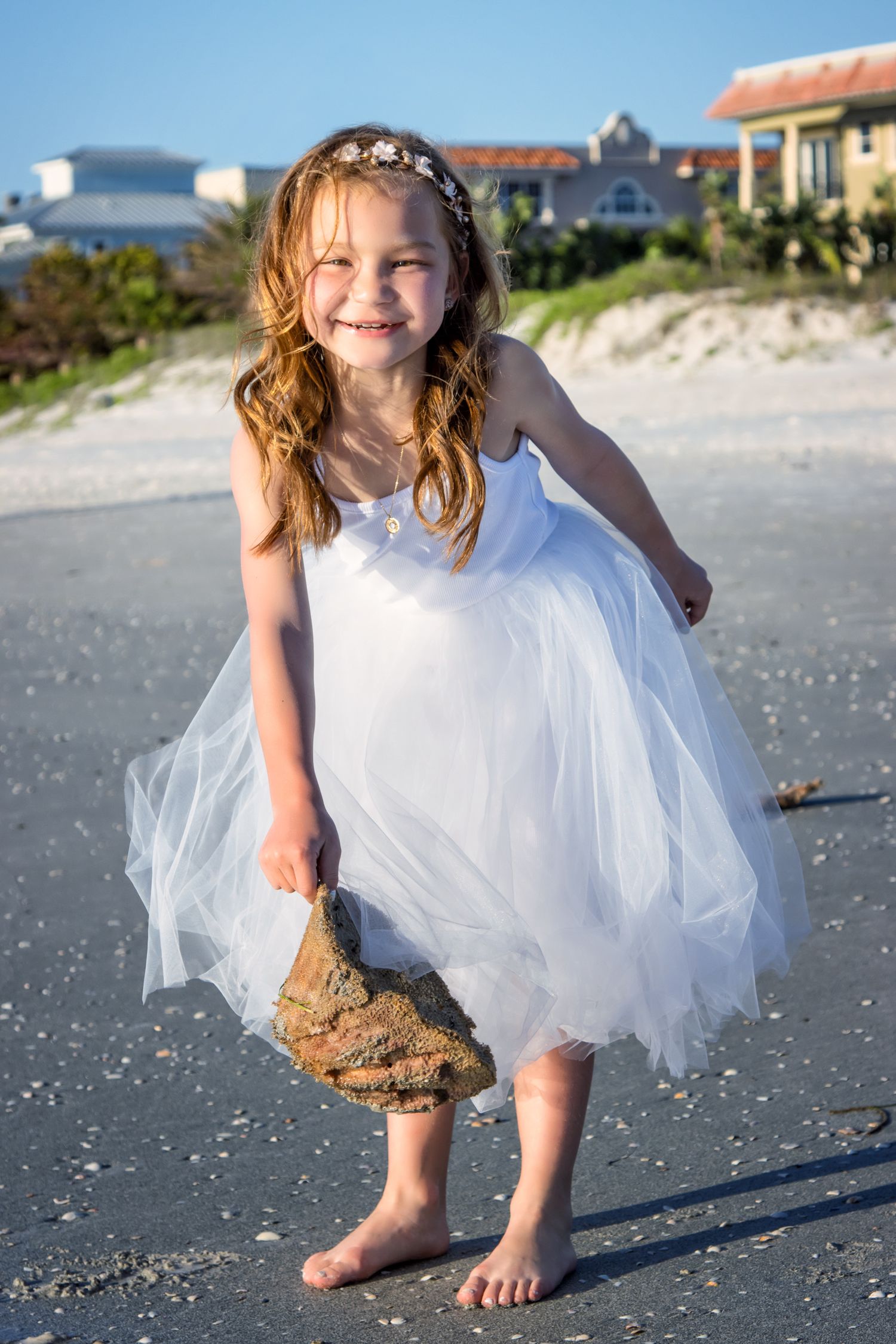 Young girl in white tulle dress and floral headband smiling on beach, holding a rock barefoot.