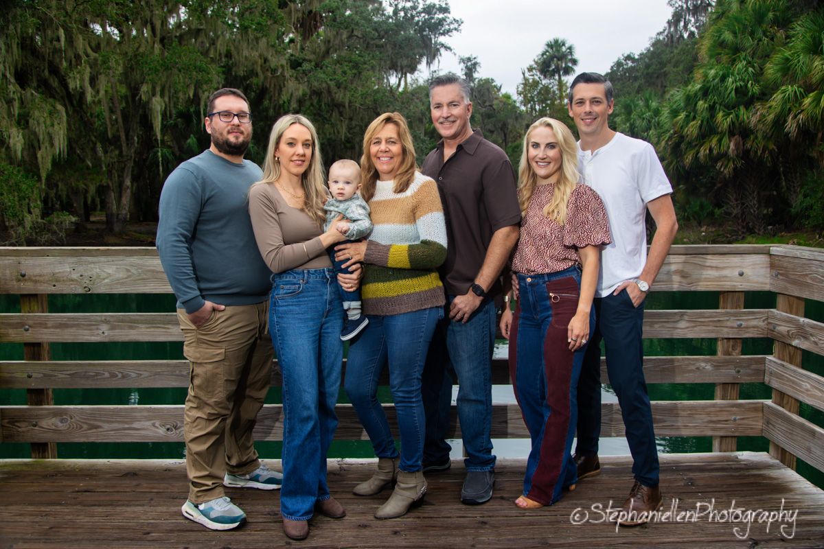 A family of seven smiles together on a wooden bridge surrounded by lush green trees in an outdoor portrait.