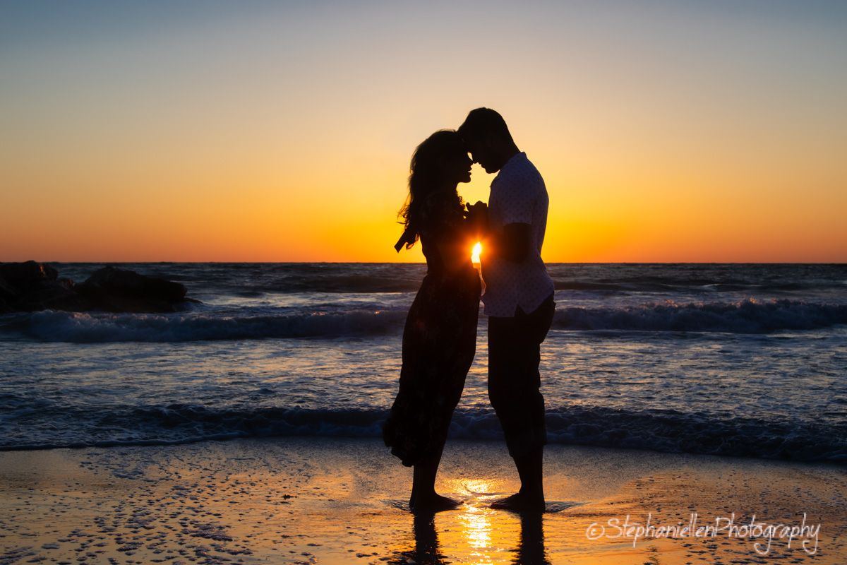 Romantic couple silhouetted kissing on the beach at sunset with golden reflections on the water.