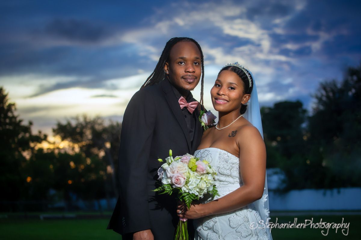 Bride and groom pose together at sunset with a colorful sky behind them.
