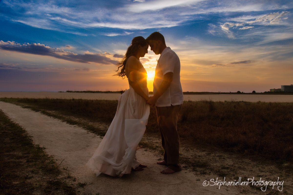 Couple sharing romantic sunset moment on a dirt path in an open field, bride in flowing white gown.