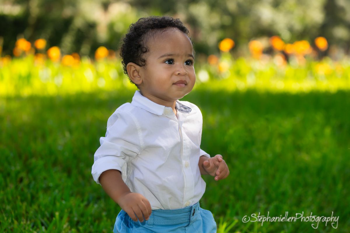 Adorable toddler boy in white shirt and blue suspenders playing on green grass with colorful flowers behind him.