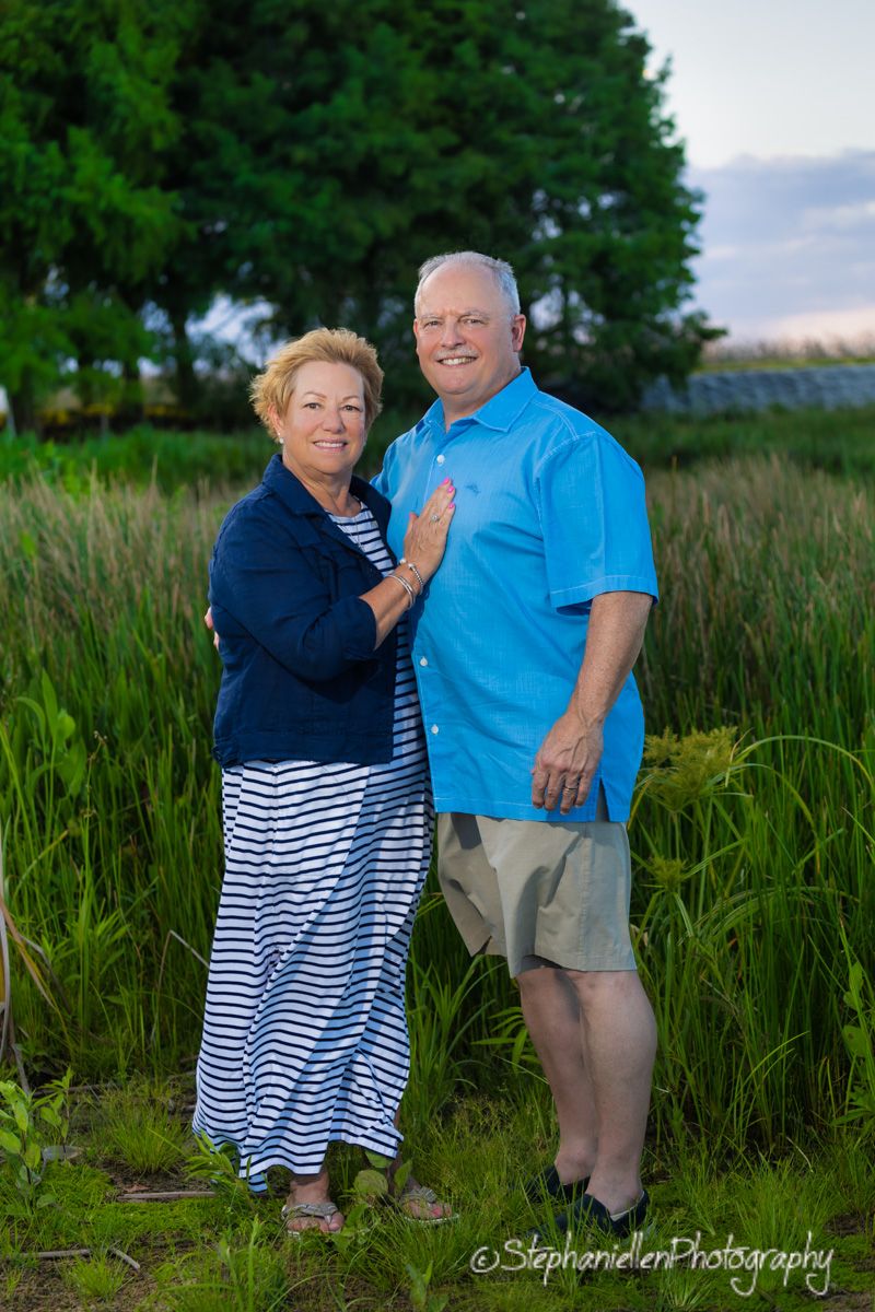 Older couple smiling and posing together in a lush green field at sunset, woman in striped dress, man in blue shirt.