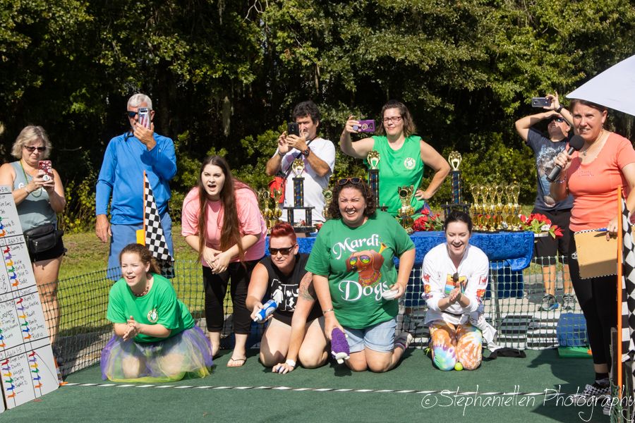 Group of people posing with a trophy and checkered flag at an outdoor racing event on a sunny day.