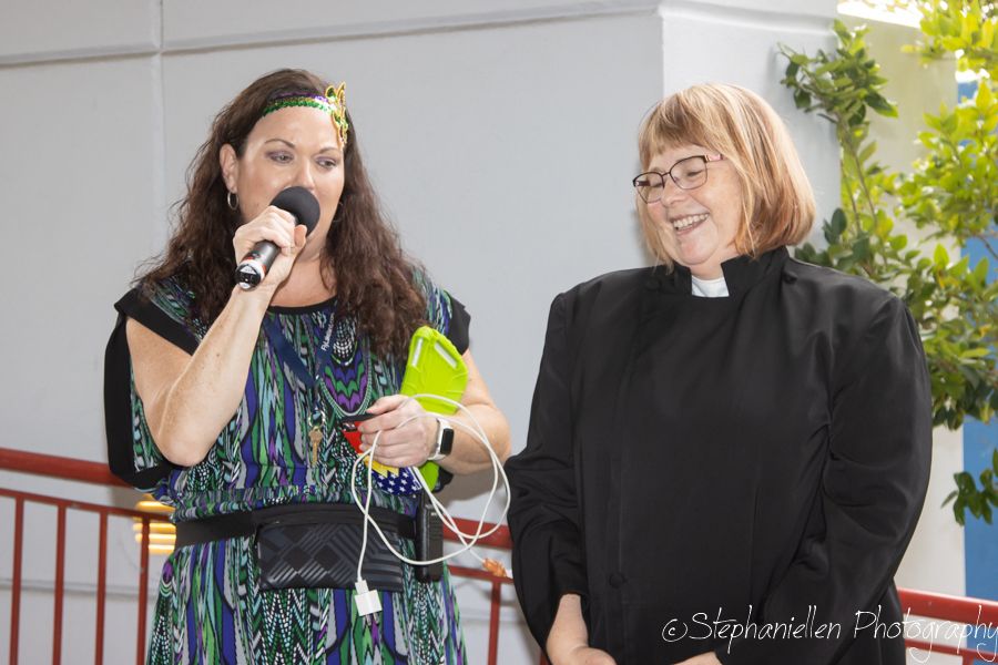 Two women stand at a podium, one speaking into a microphone wearing a colorful dress, the other in black clergy attire.