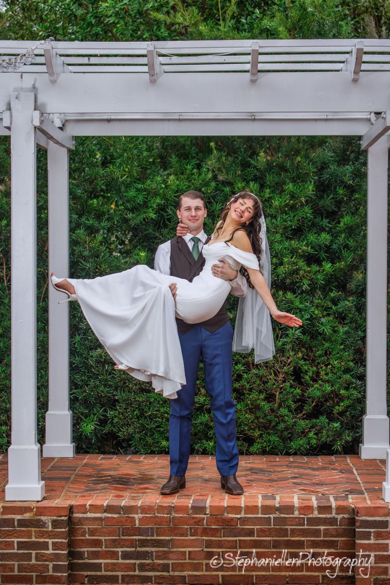 Joyful groom lifting bride in white dress under white pergola with green hedge backdrop.