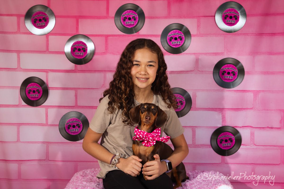 A smiling girl holds a small dachshund with a pink bow in front of a pink brick wall with vinyl record decorations.