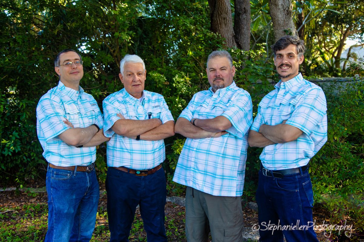 Four men in matching plaid shirts stand with arms crossed outdoors, posing for a group portrait photo.
