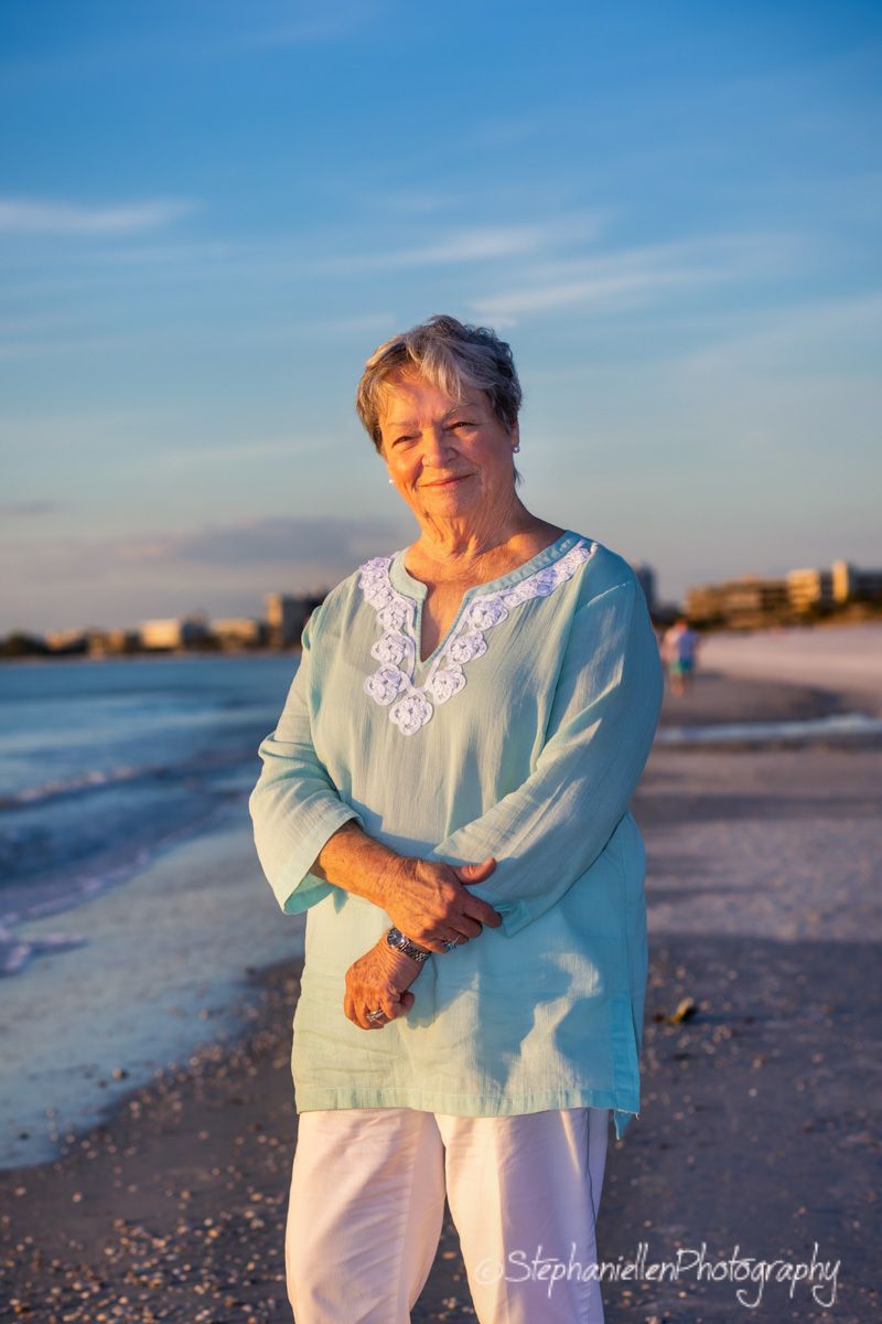 Smiling older woman in teal tunic and white pants posing on a sunny beach at golden hour.