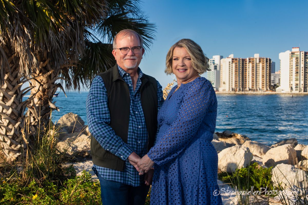Couple posing together near rocky shoreline with ocean and high-rise buildings in background on sunny day.