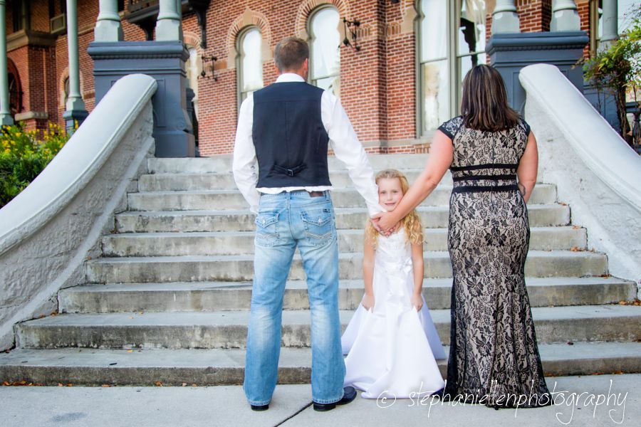 Family of three holding hands on steps of historic brick building, viewed from behind.