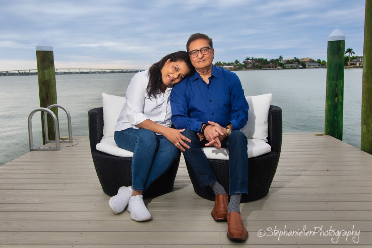 Couple sitting together on a waterfront dock chair, smiling, with ocean and dock pilings in the background.