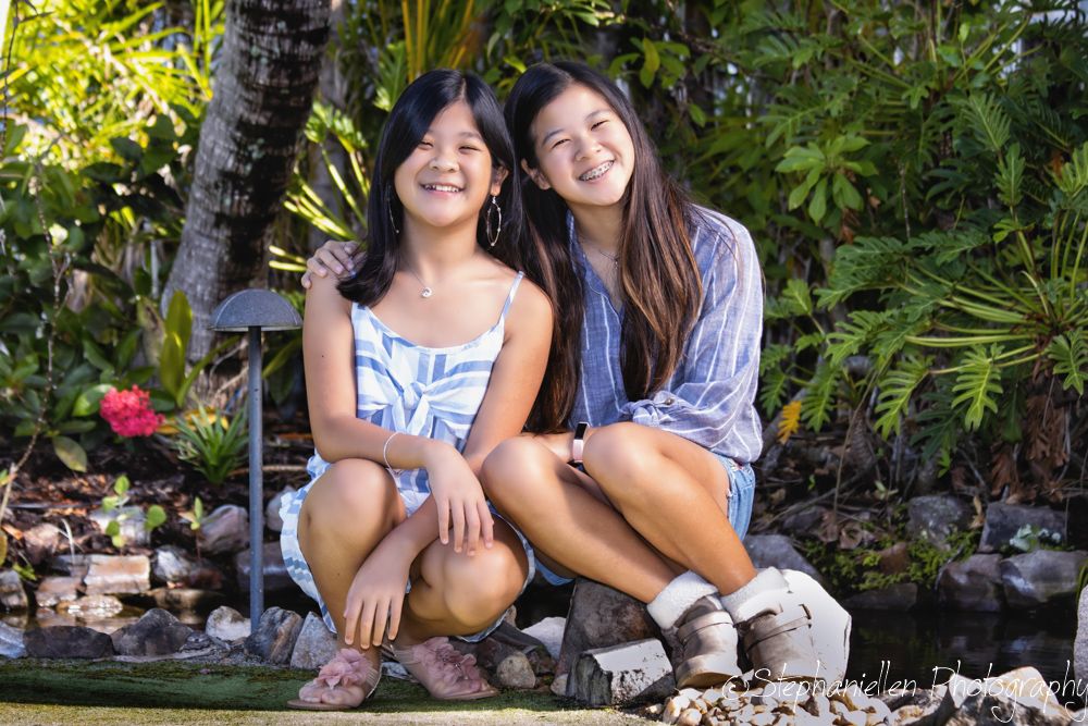 Two smiling young women pose together in a lush tropical garden near a pond.