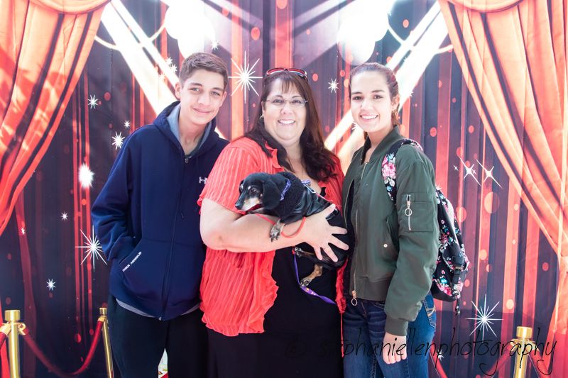 Woman holding a black dachshund poses with two teens in front of a red curtain backdrop.