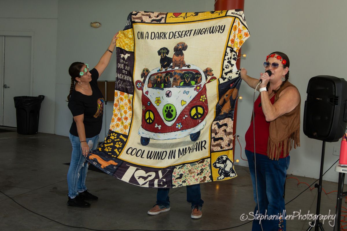 Two women display a colorful hippie-themed quilt with a VW bus design at an indoor event.