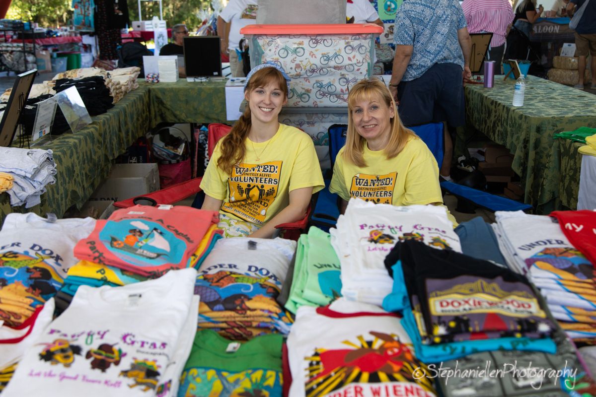 Two smiling women in yellow shirts sell colorful t-shirts at an outdoor market booth.