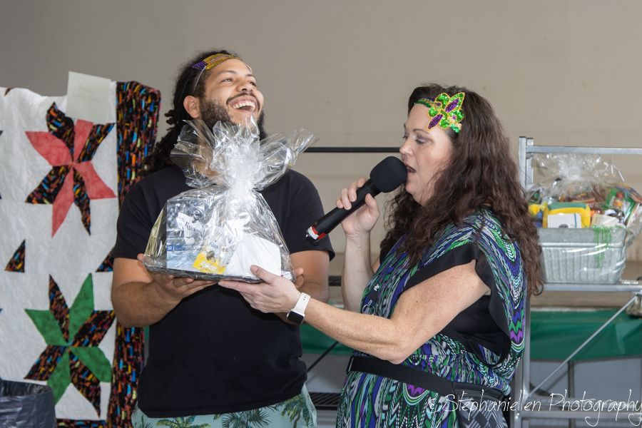 A laughing man holds a gift basket while a woman speaks into a microphone at an indoor event.