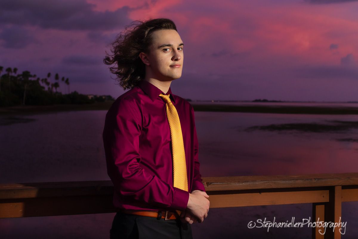 Young man in maroon shirt poses on bridge at stunning purple sunset waterfront.