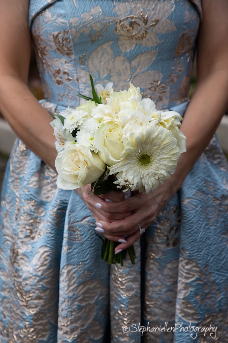 Bride holds elegant white bouquet of roses and gerbera daisies against blue brocade gown.