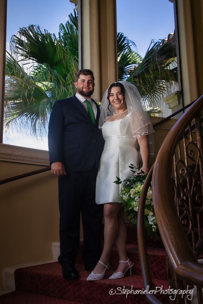 Bride and groom posing on a staircase by a large window with tropical palm trees in background.