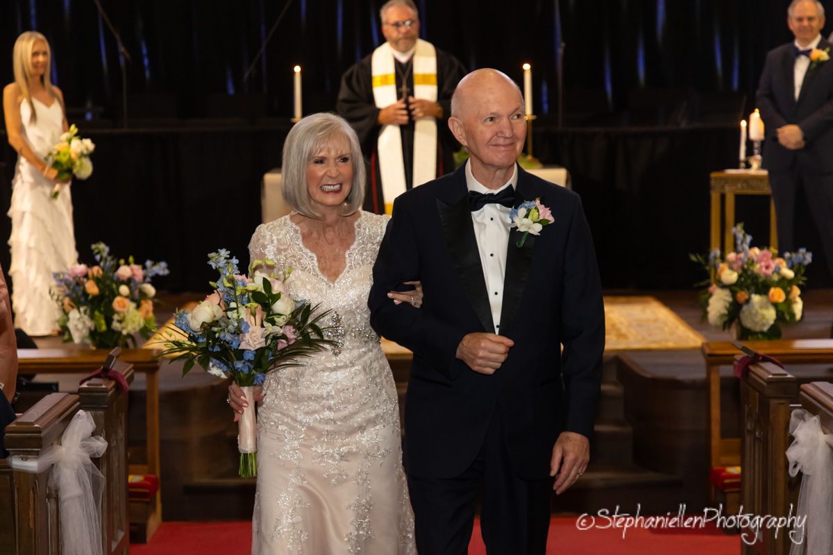 Older couple walks down church aisle smiling after wedding ceremony with floral arrangements.