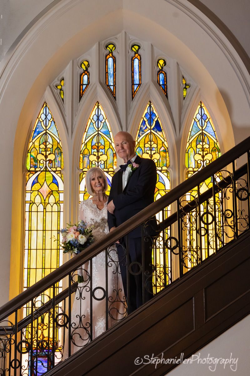 An elegant older couple poses on a grand staircase before stunning stained glass windows in a historic church.