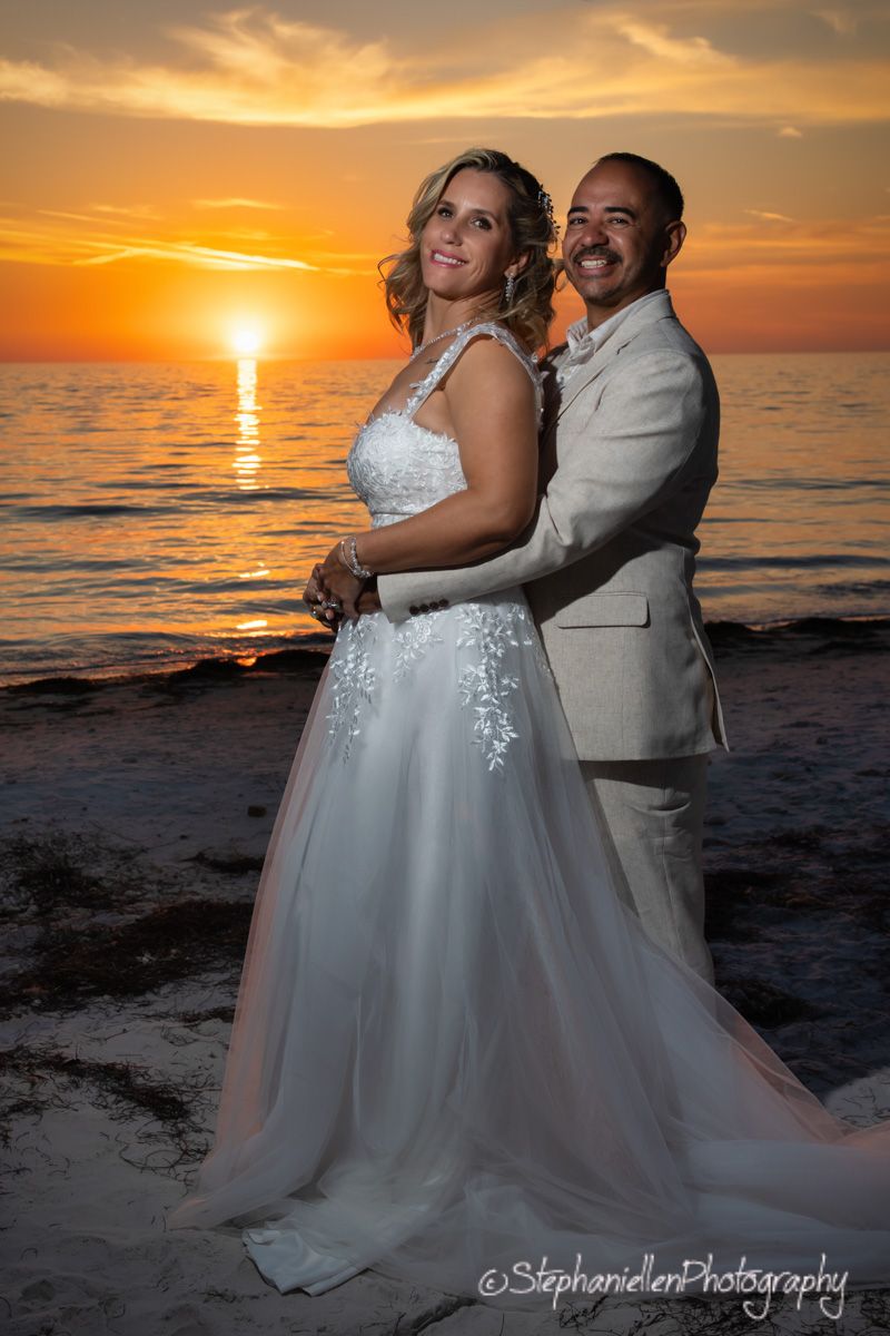 Bride and groom embrace on beach with vibrant orange sunset over ocean.