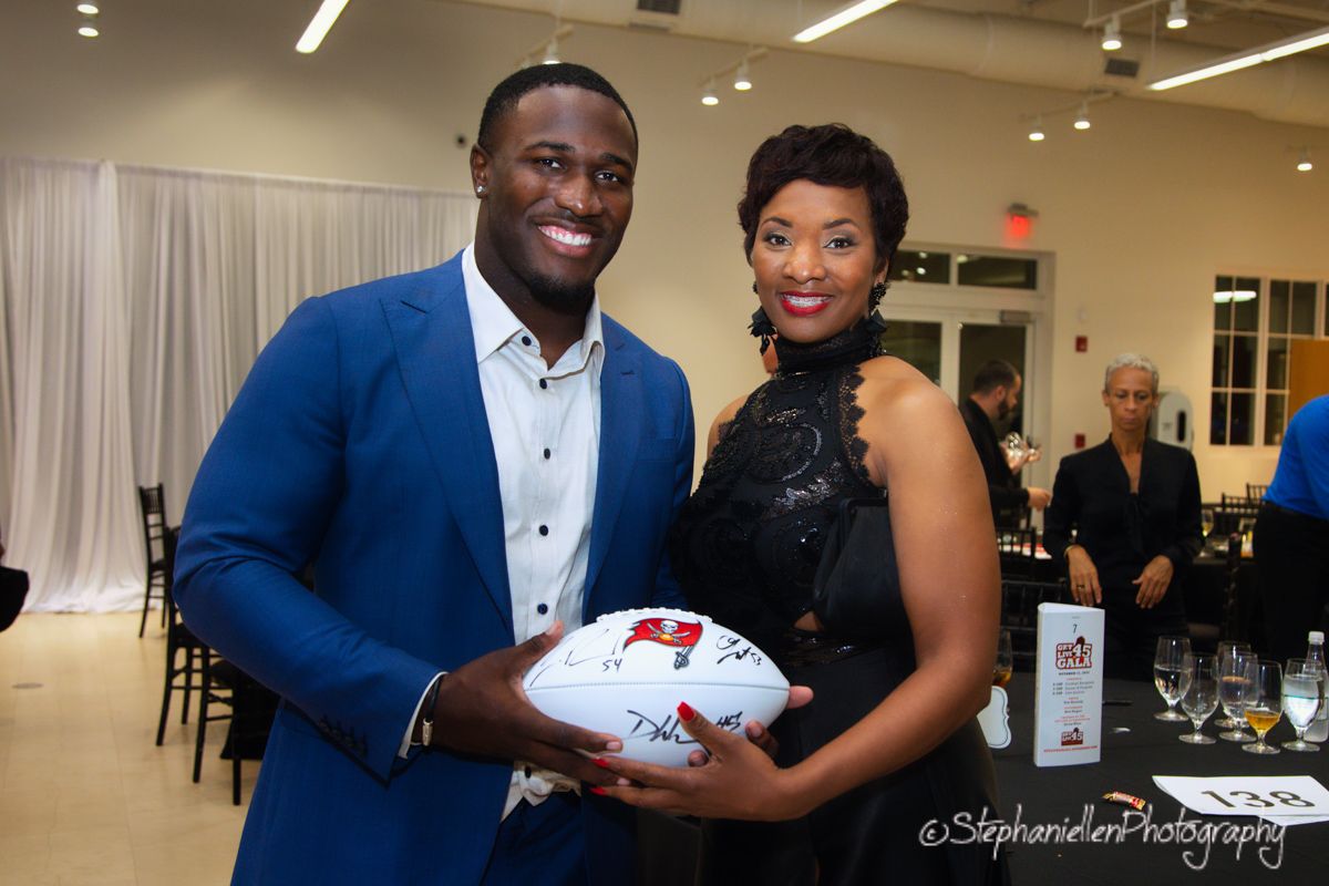 Man in blue suit and woman in black dress posing with signed Tampa Bay Buccaneers football.