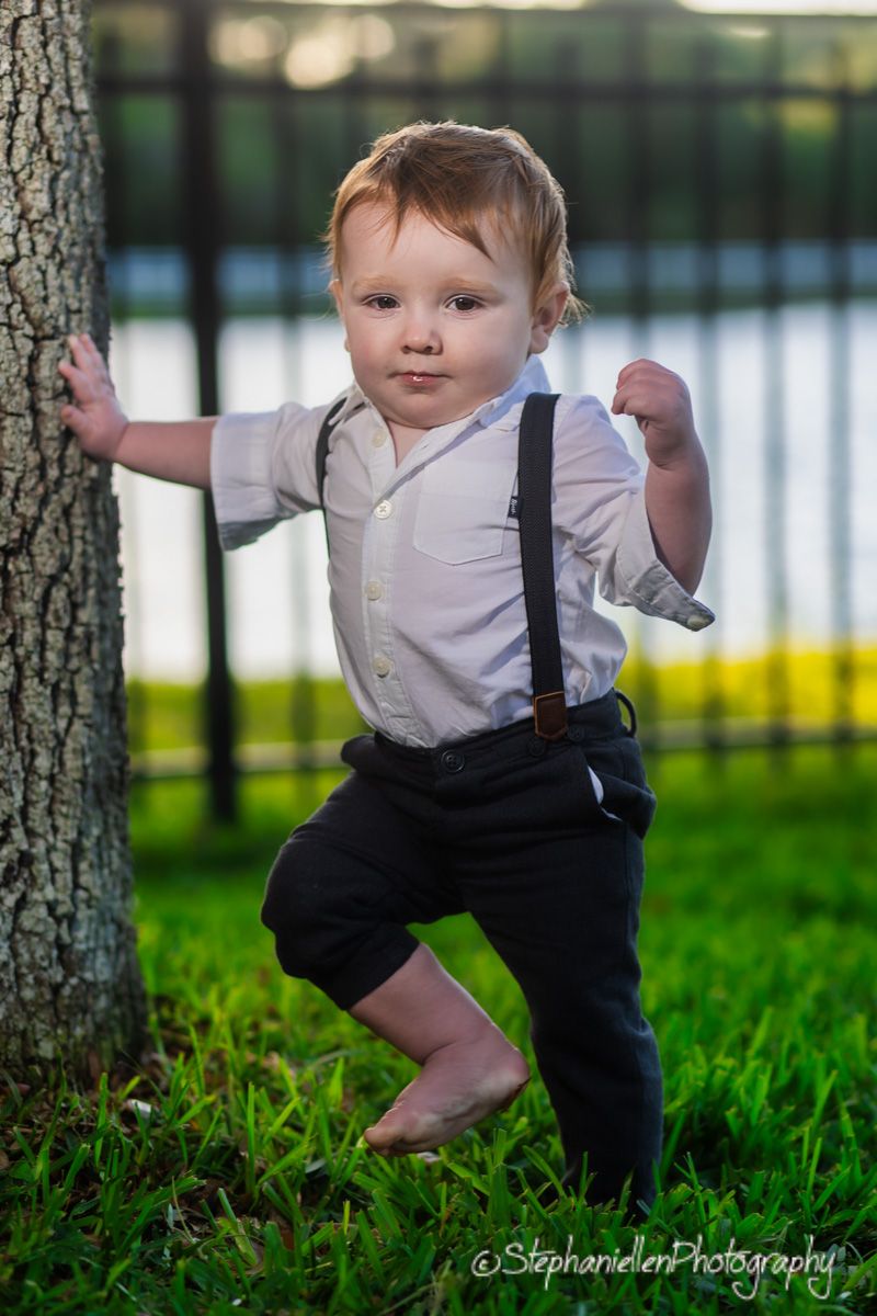 Adorable toddler boy in white shirt with suspenders and black pants standing barefoot on green grass by a tree.