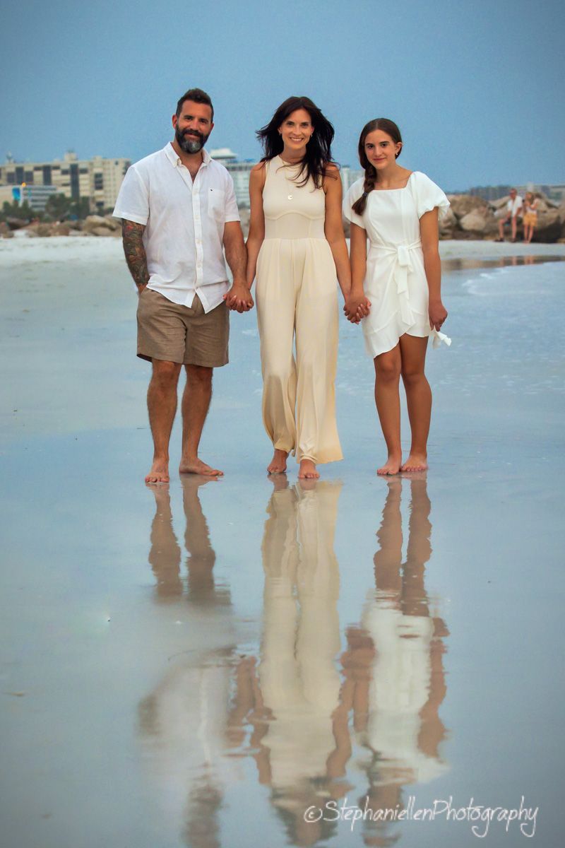 Family of three posing on a reflective beach shore, wearing white and cream outfits, with city skyline in background.