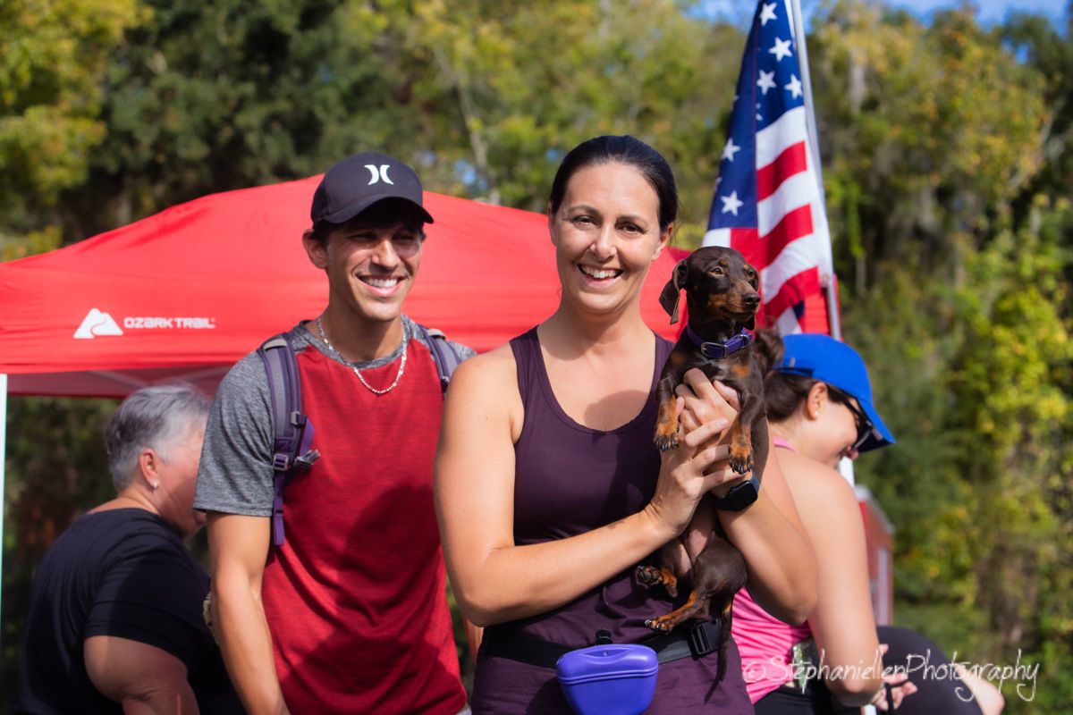 Two smiling people holding a small dog at an outdoor event with an American flag and red tent in background.